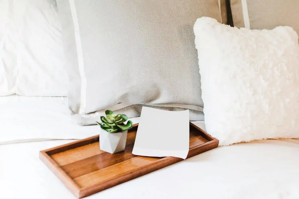 Bett mit weißen Laken. Darauf liegt ein Holzbrett mit einer Pflanze und Papier.